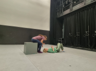 One performer sitting on a box looking at the palm of her hand, another lying on the floor directly below looking up at her.