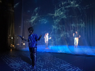 Lit stage space, actor standing rehearsing with script, two dancers standing up stage.